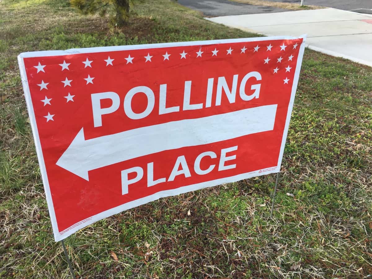 Polling place sign placed in a grassy area wit a sidewalk in the background.