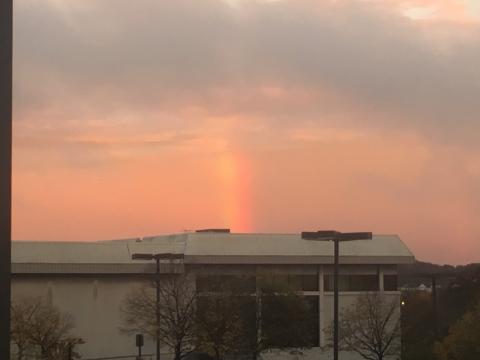 A picture of a landscape. In the foreground is a nondescript building, and some street lights. Above the buildings the sky fades from pink to dark blue-gray. In the middle of the sky, below the blue-gray clouds is a bright beam of a rainbow.