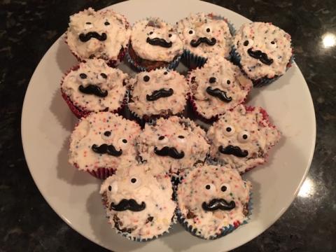 A plate containing a dozen cupcakes. The cupcakes are covered in fun-fetti icing, and decorated with candy eyeballs and moustaches to look like faces. Each cake's eyes and faces look sightly different, giving each one a unique expression, while appearing to look in different directions.